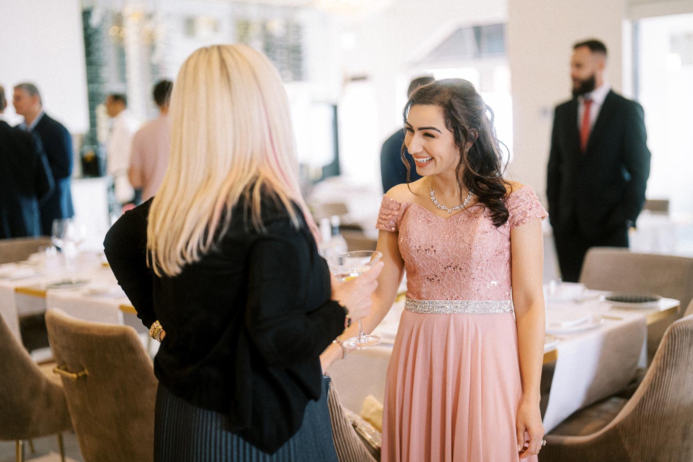 Two women in elegant attire smiling and conversing at an indoor social event, with seated guests and a stylishly set table