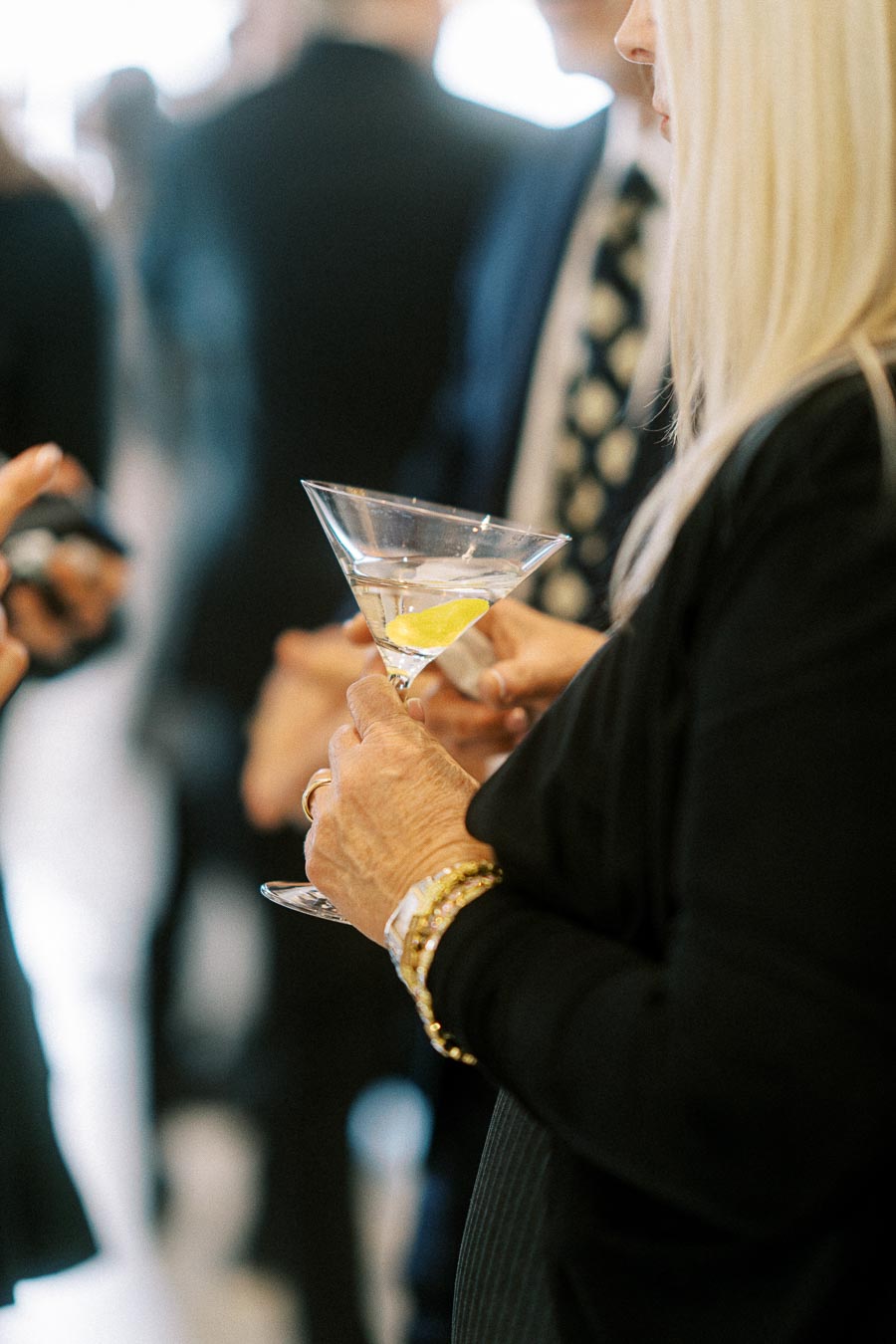 A person holding a martini glass with a lemon twist at a social gathering, wearing gold bracelets and black outfit.