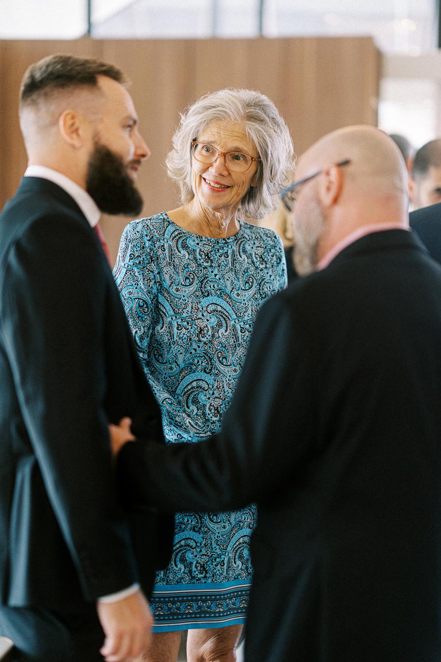 Senior woman in a blue patterned dress smiling and interacting with two men in suits at a social event indoors.