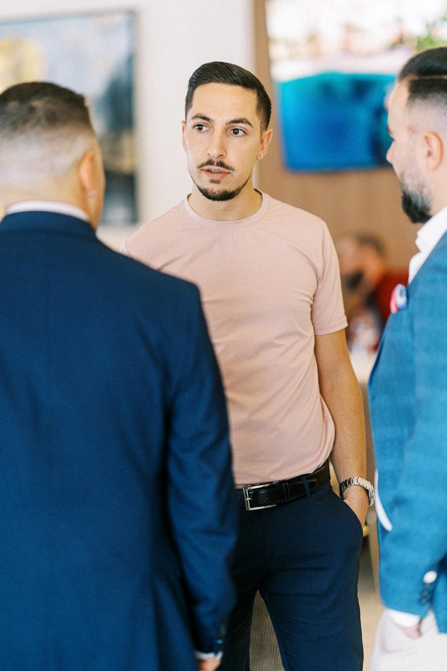 A man in a pink t-shirt engaging in a conversation with two men in suits, set in a professional indoor environment.
