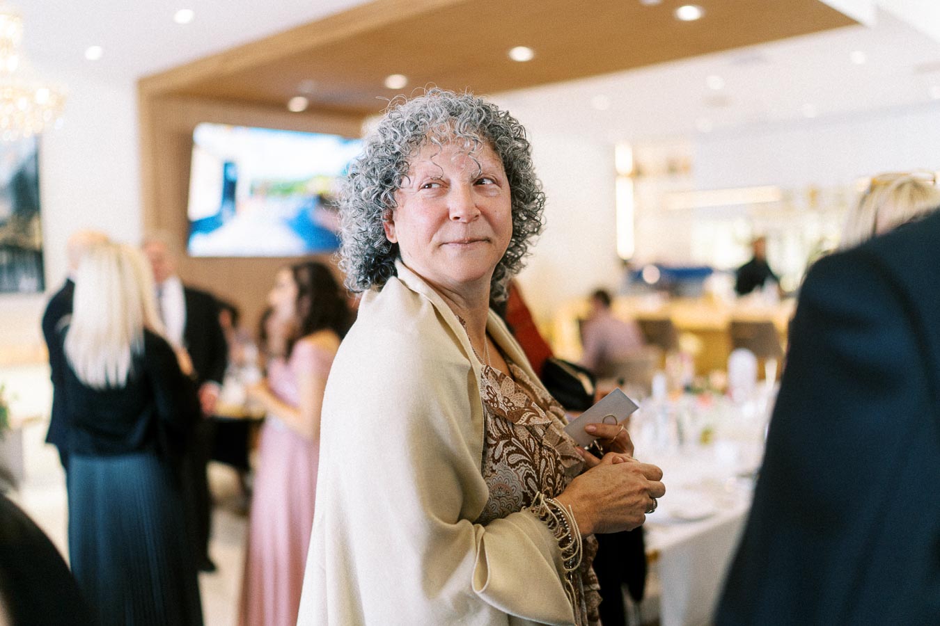 Elderly woman with curly gray hair attending a social event, dressed in a patterned outfit with a beige shawl, surrounded by