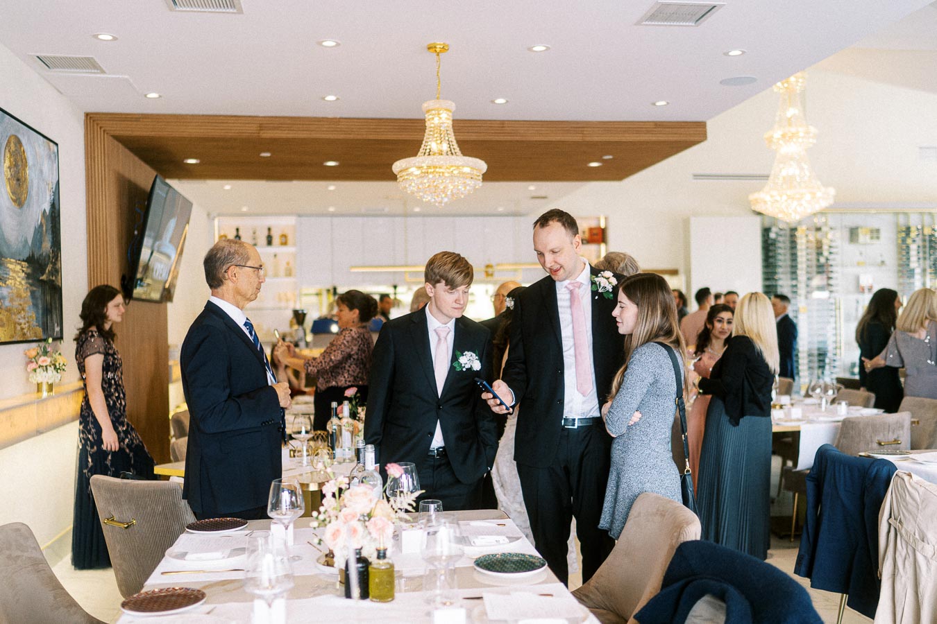 A group of well-dressed people socializing in an elegant restaurant setting with chandeliers and floral decor, suggesting a