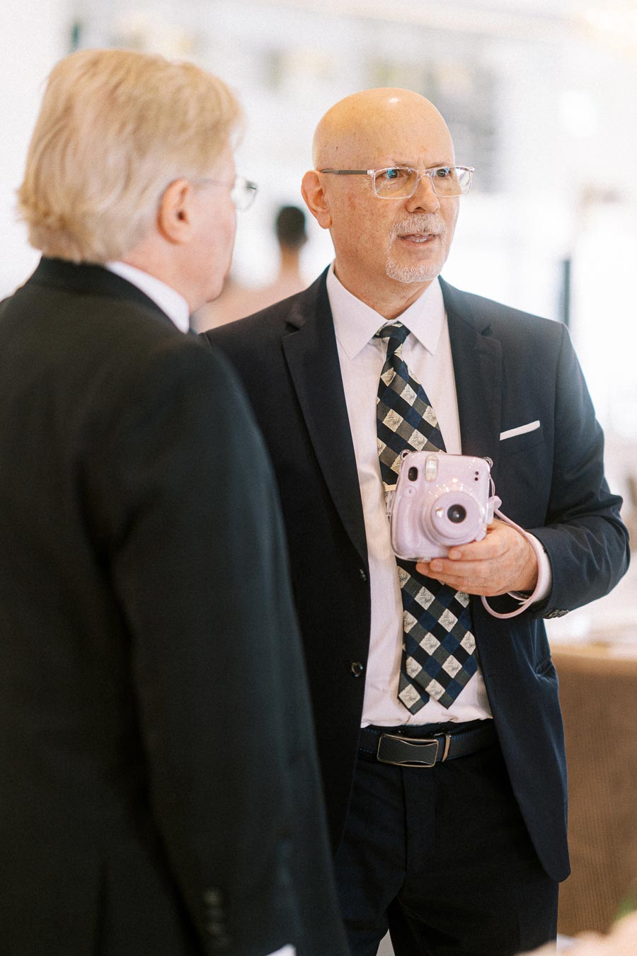 Two professionally dressed men engaged in conversation, with one holding a pink instant camera at a business event.