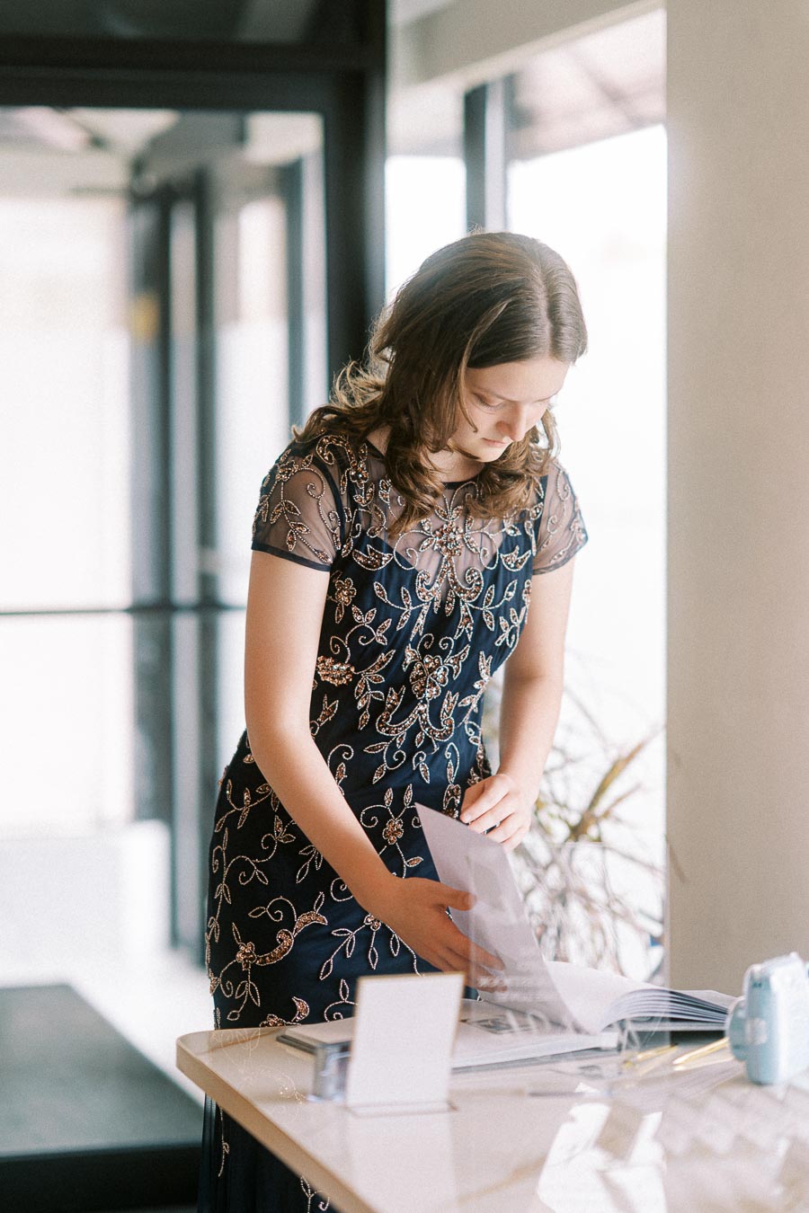 Young woman in an elegant navy blue dress with intricate floral embroidery, standing by a bright window and browsing through
