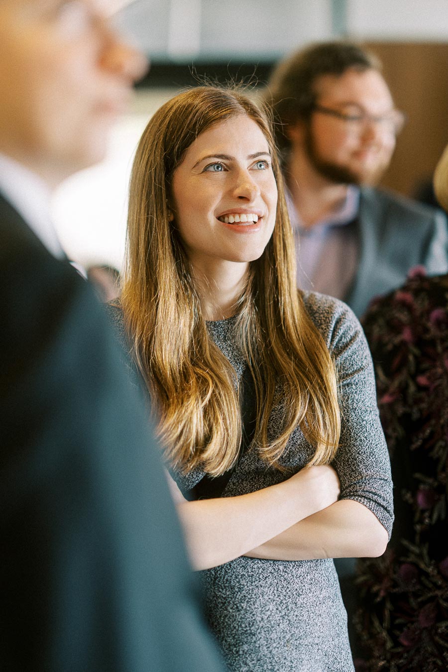 Young woman with long hair smiling at a social event, surrounded by people in blurred background.