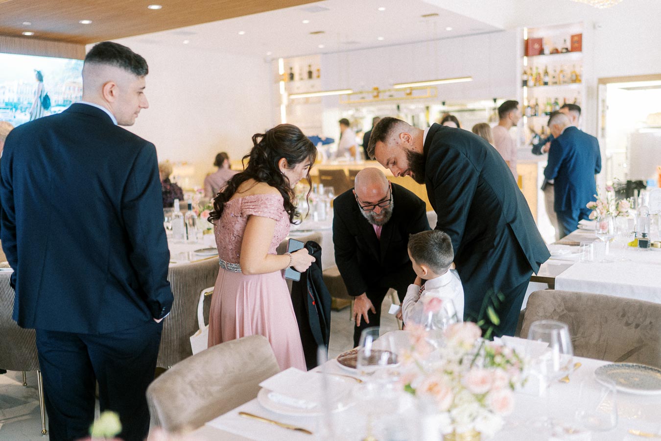 A group of well-dressed guests interact at a formal event in a beautifully decorated venue, featuring elegantly set tables