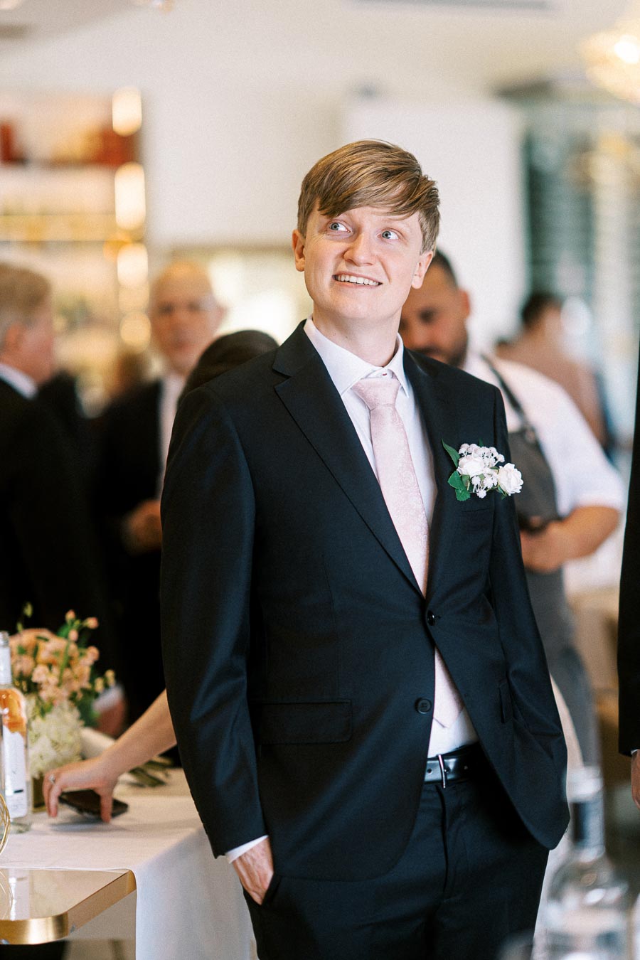 A well-dressed man in a black suit and pink tie smiles at a formal event, surrounded by elegantly dressed guests in a