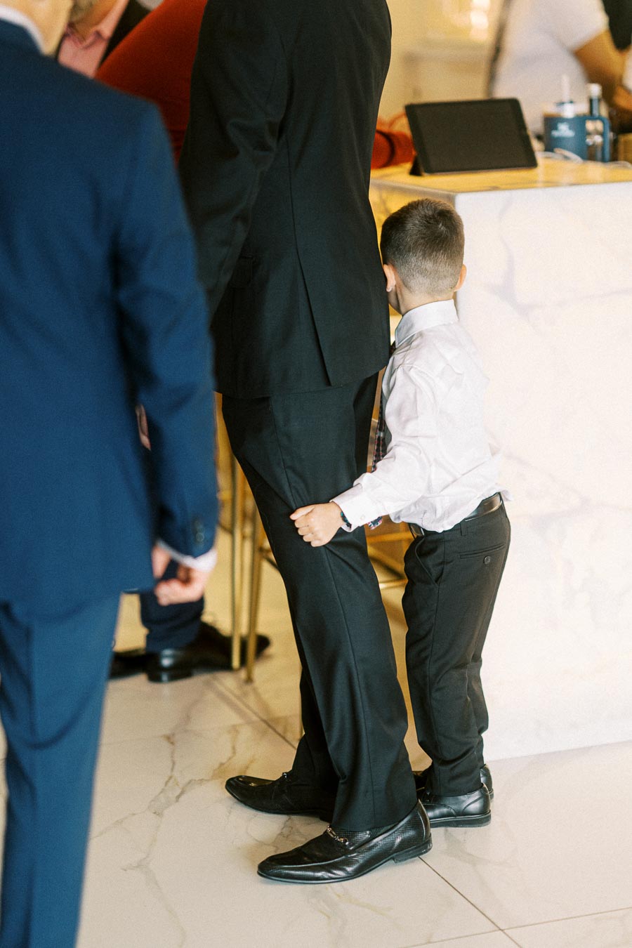 A young boy in a formal outfit playfully peeks out from behind an adult at a social event, highlighting a family gathering