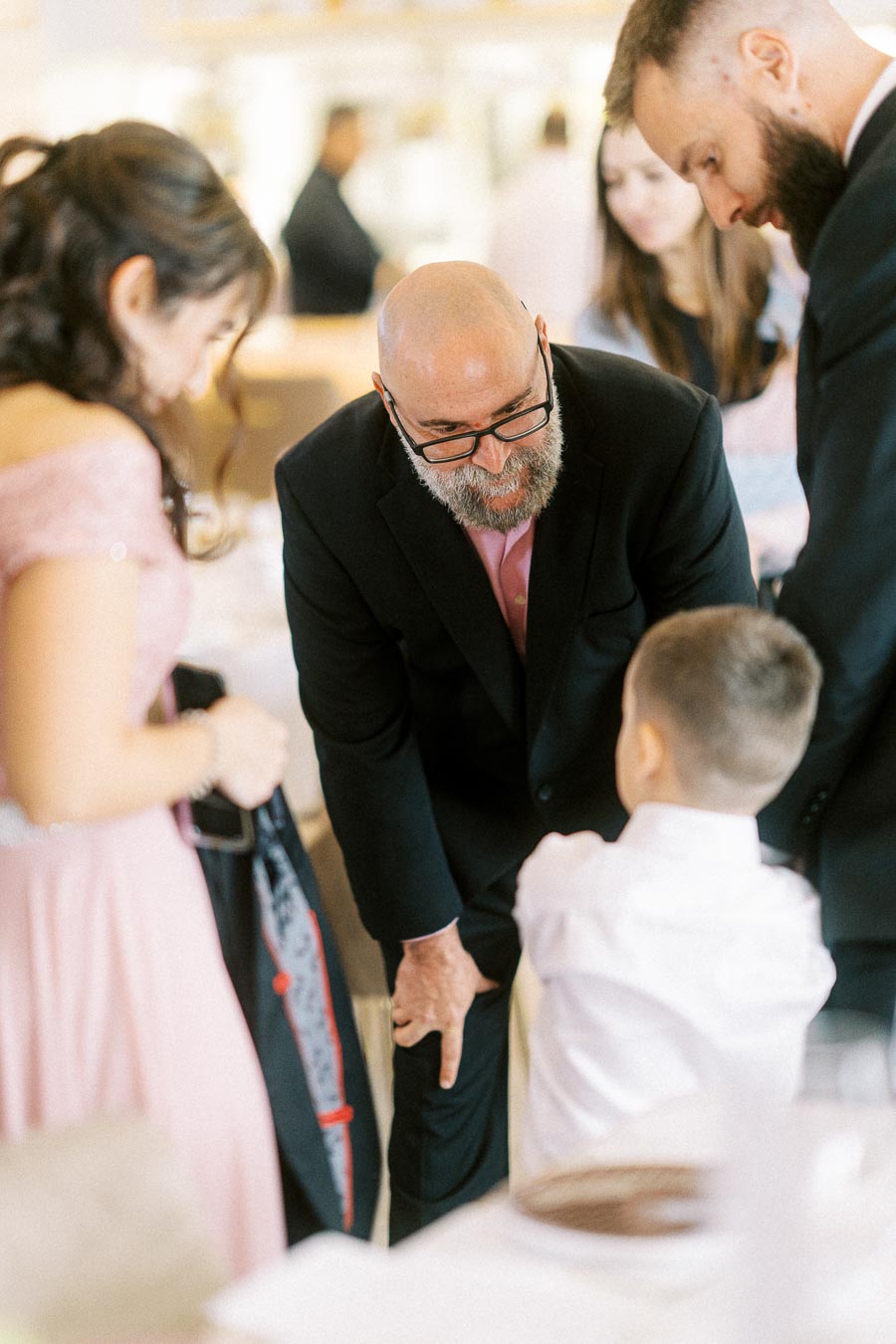 A group of people at a formal event, including a man in a suit crouching to speak with a young boy, surrounded by other