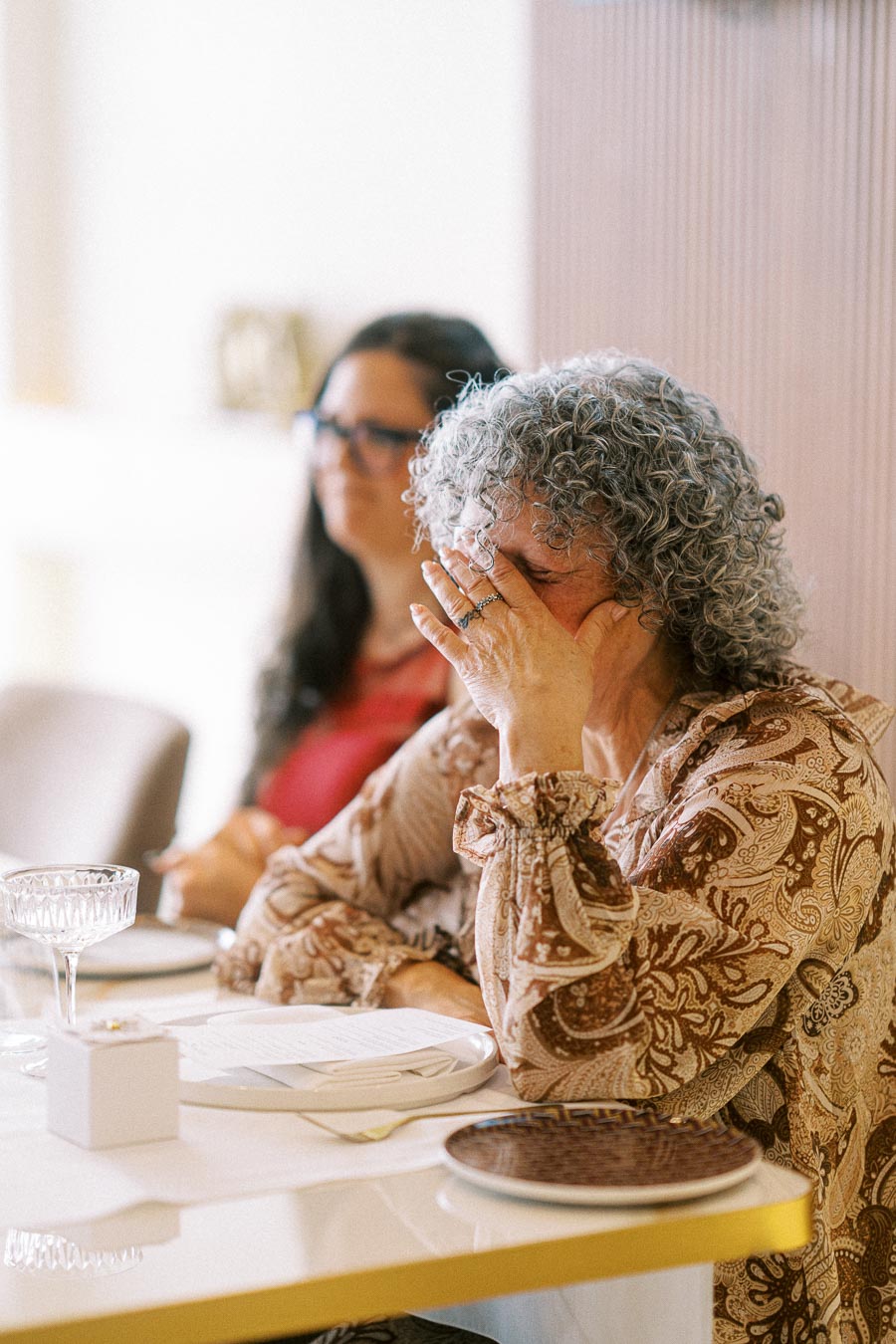 Elderly woman at a dining table covering her face with her hand, expressing emotion, while another person sits in the