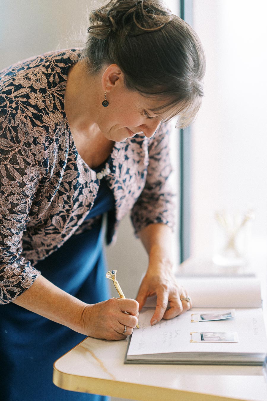 A woman signing a guestbook at a wedding while wearing a blue dress and a floral-patterned jacket, using a decorative pen.