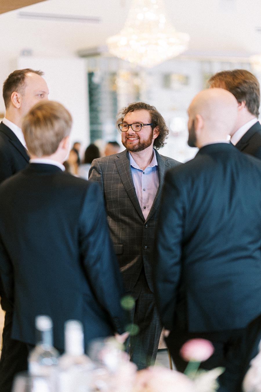 A group of men in suits engaged in conversation at a formal event, with one smiling man in a checked suit in focus,