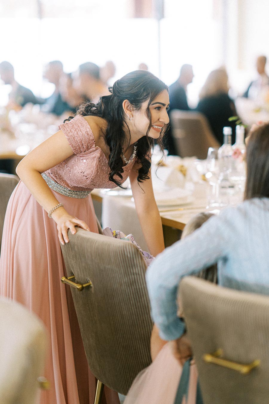A young woman in an elegant pink dress with lace details leans over to chat with guests at an indoor social gathering. The