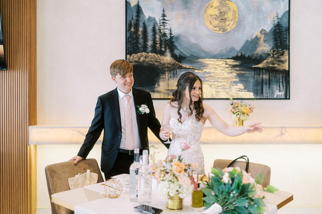 A bride and groom in formal attire smiling and interacting at a decorated table with floral arrangements, under a landscape