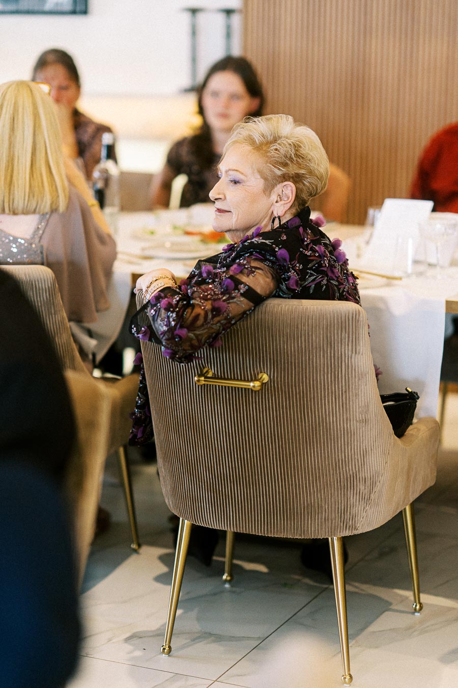 Elderly woman in a floral blouse sitting at a dining table with others at a formal event, focused and engaged in
