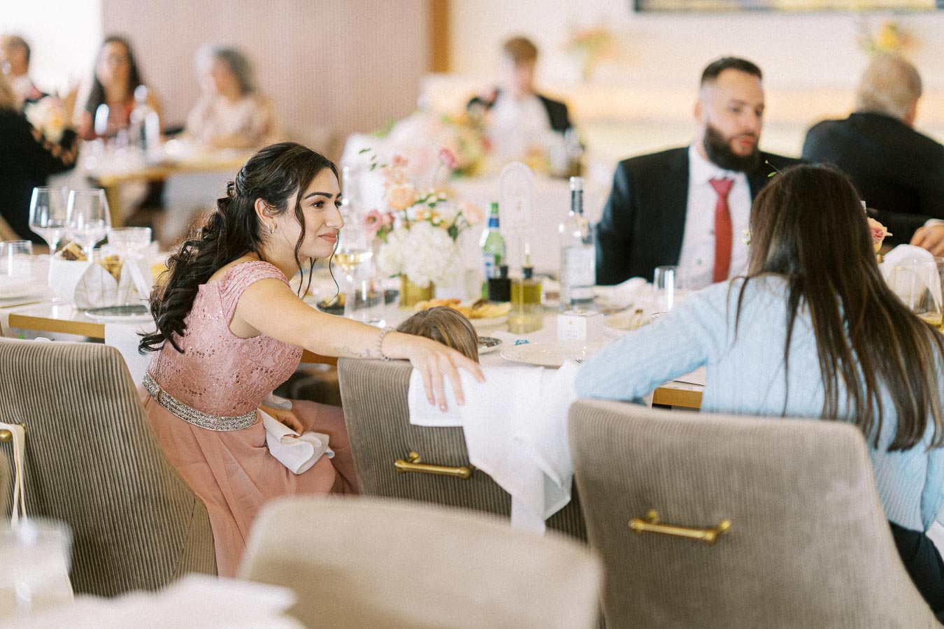 A woman in a pink dress reaching across a dining table at a formal event, with elegantly set tables and guests in the