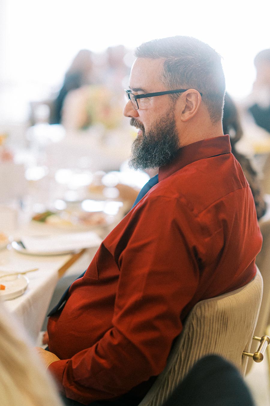 A person with a beard and glasses wearing a red shirt sitting at a dining table in a bright, softly blurred setting.