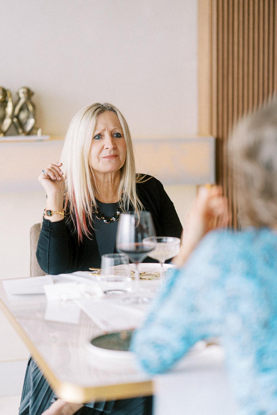 A woman with long blonde hair engaged in a conversation at a dining table, with wine glasses and tableware, creating an