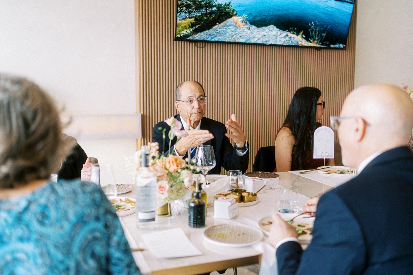 A group of people in formal attire engaged in conversation around a dining table with elegant settings and floral