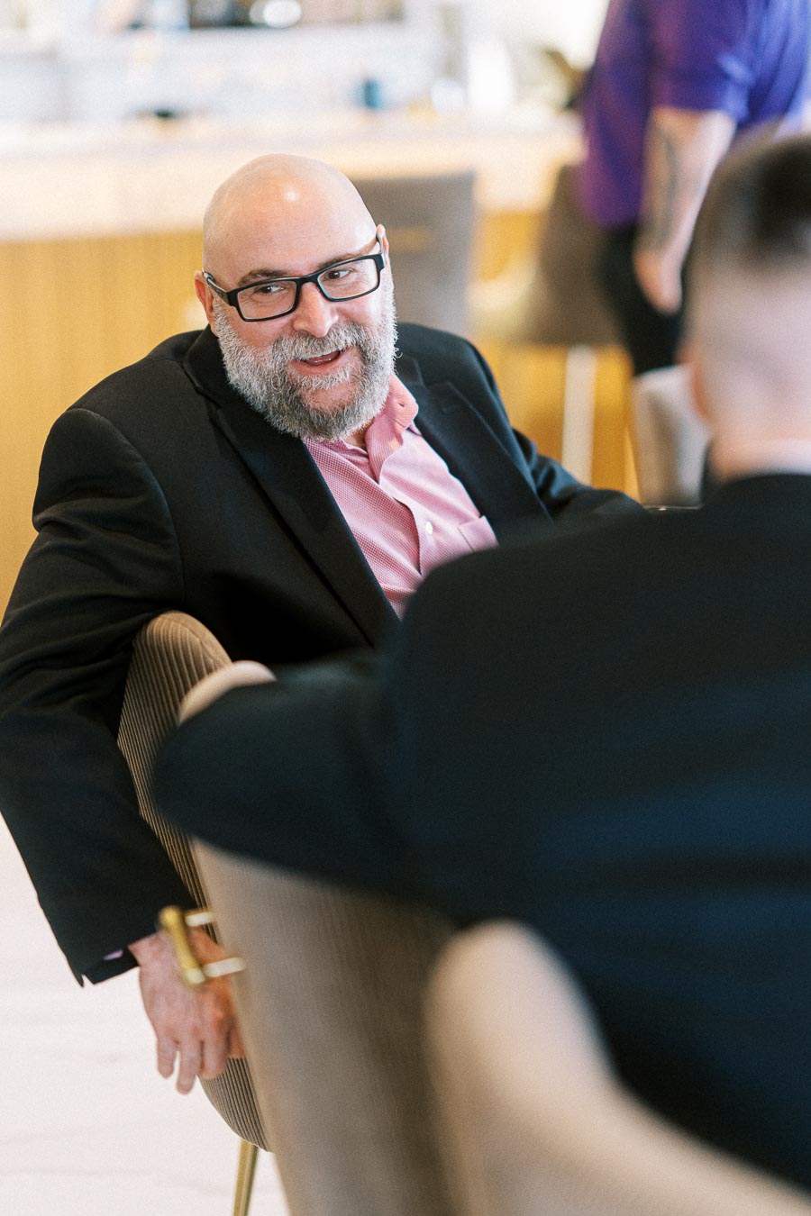 A bearded man in glasses, wearing a black blazer and pink shirt, smiling and engaged in conversation at a business meeting