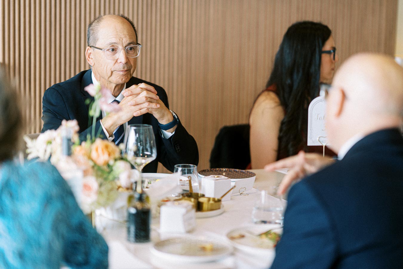 Elderly man in a suit sitting at an elegant table with floral decorations and people engaged in conversation at a formal