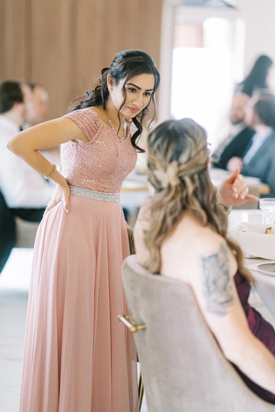 Woman in a pink lace dress talking to a seated guest at an indoor event.