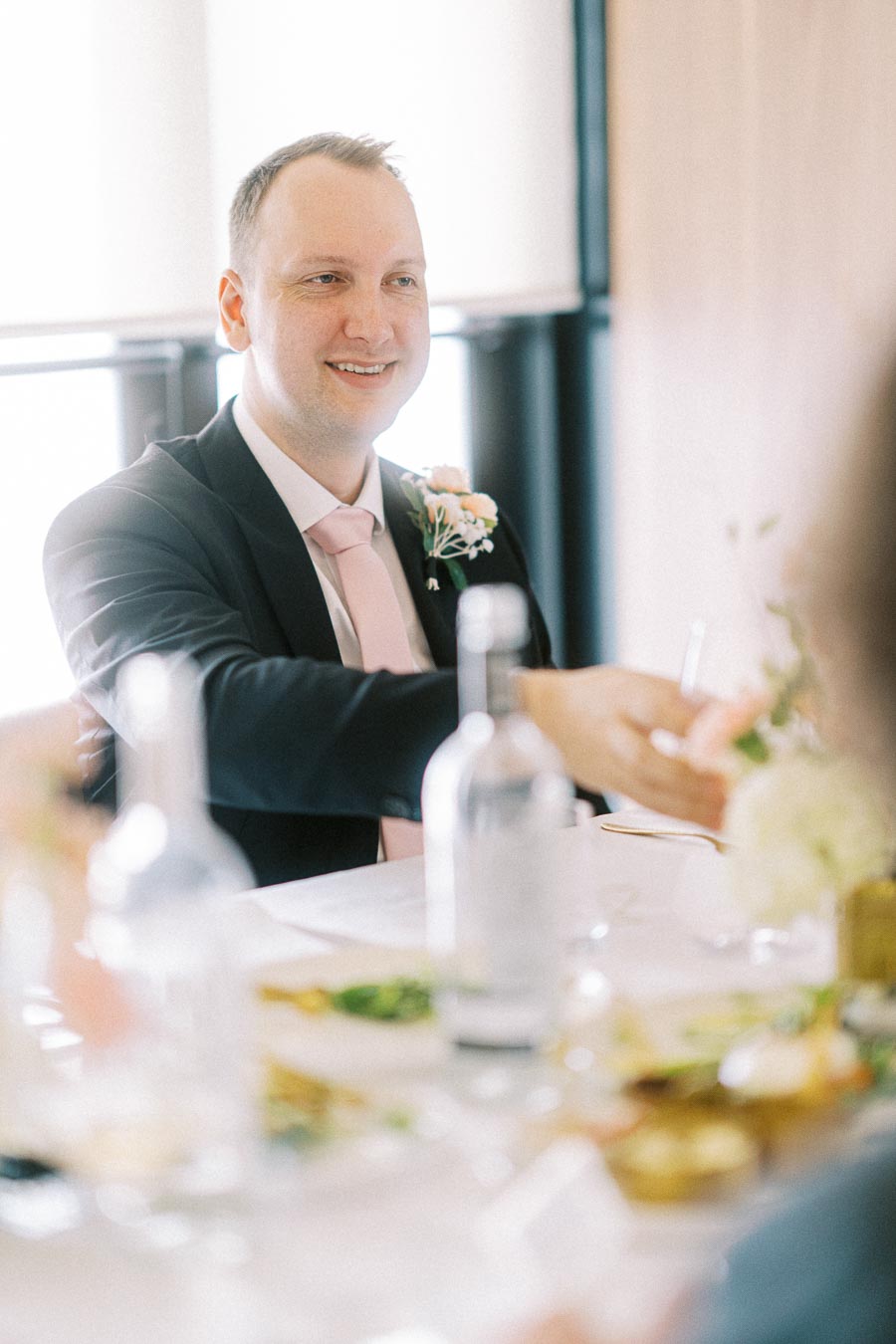 A man in a suit smiles at a formal table setting, featuring white linens, glassware, and floral decorations, in a bright,