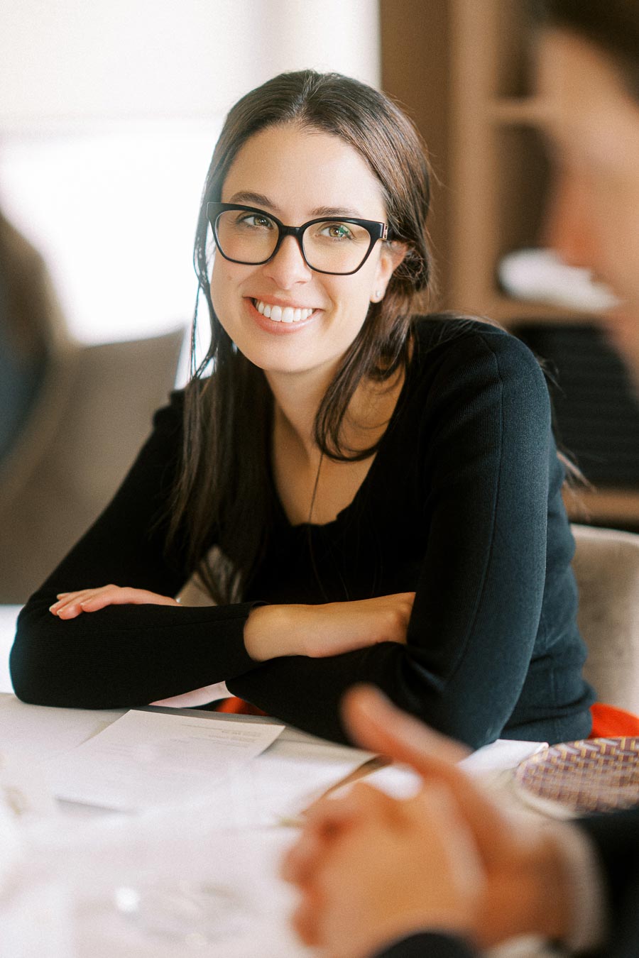 Woman with glasses smiling at a table, engaged in conversation.
