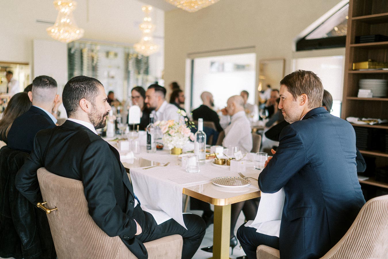 Elegant restaurant setting with well-dressed guests seated at a dining table, engaging in conversation. The table is adorned