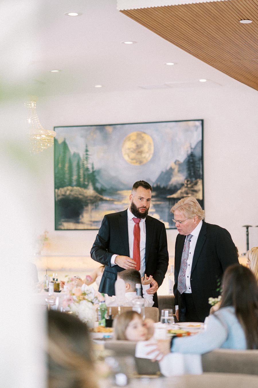A busy banquet hall with two men in suits having a conversation near a beautifully set table, surrounded by guests and