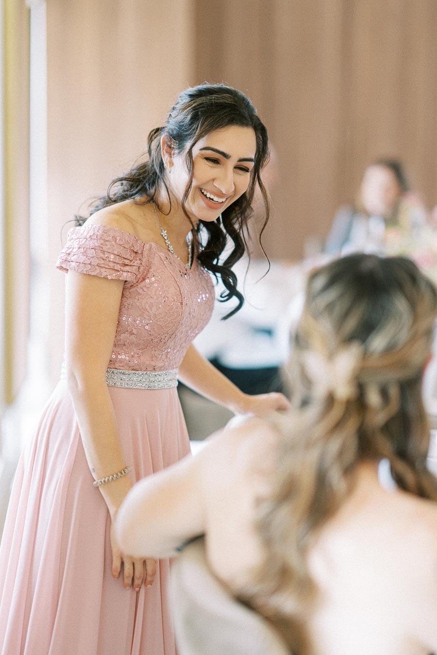 A woman in a pink lace gown smiles at another seated woman in a softly lit, elegant indoor setting.
