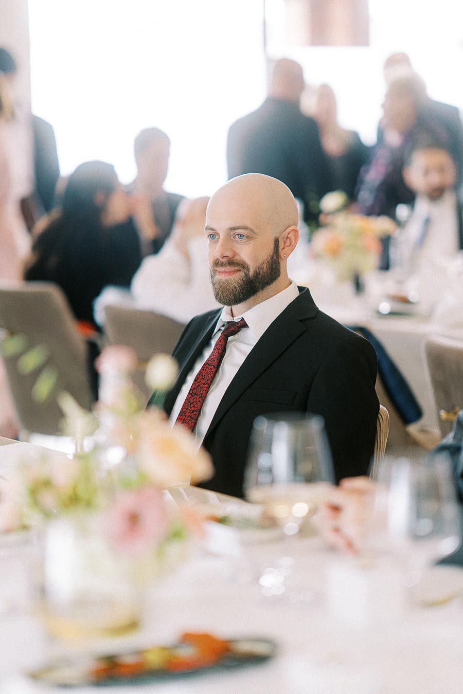 A man with a beard and a bald head wearing a black suit and red tie seated at a beautifully decorated table during an