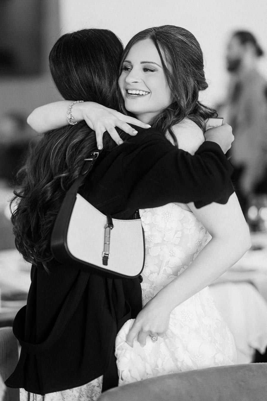 A bride joyfully embraces a friend at a wedding reception, showcasing her lace dress and radiant smile in a heartfelt black
