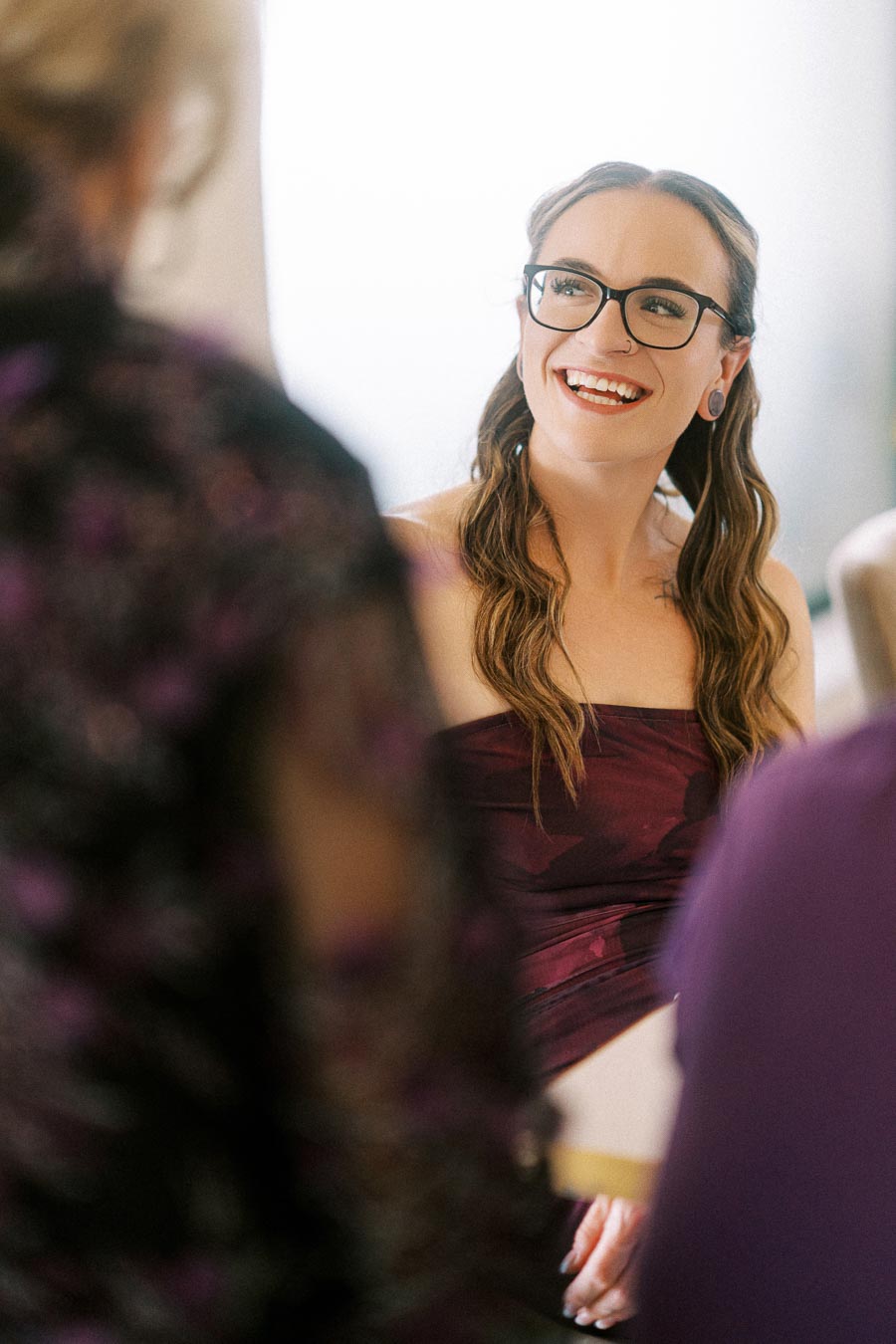 Young woman with glasses smiling in a formal setting, wearing a strapless purple dress while engaging in a conversation.