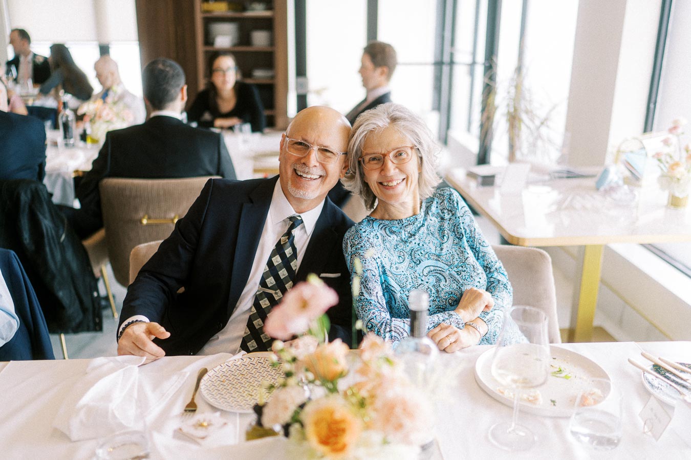 Couple smiling at restaurant table during daytime event, surrounded by floral arrangements and elegantly set dishes in a