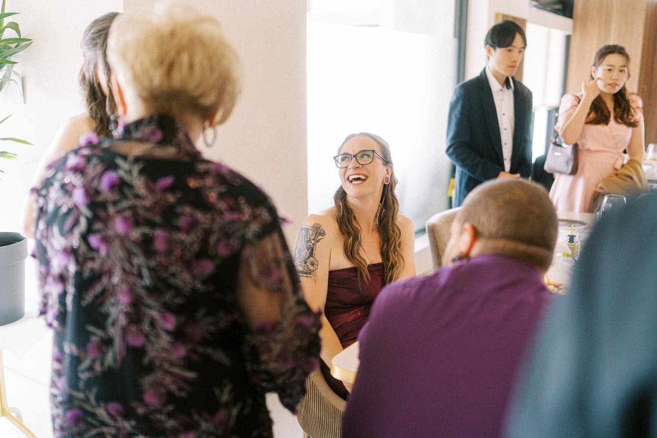 A joyful gathering at a social event, featuring a smiling woman with glasses and a tattoo, engaged in conversation with