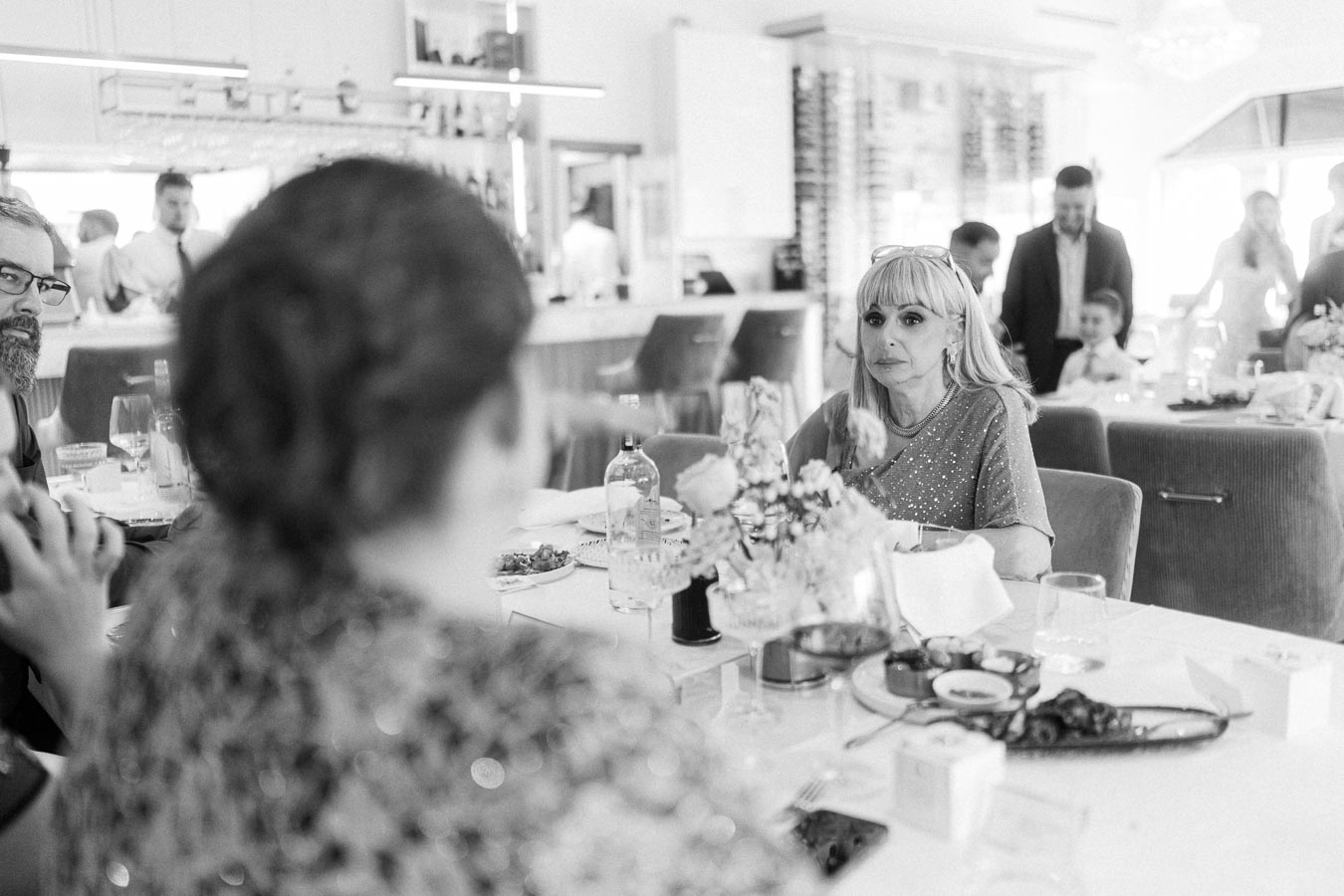 Black and white photo of a social gathering in a restaurant, featuring elegantly dressed guests sitting at a table adorned