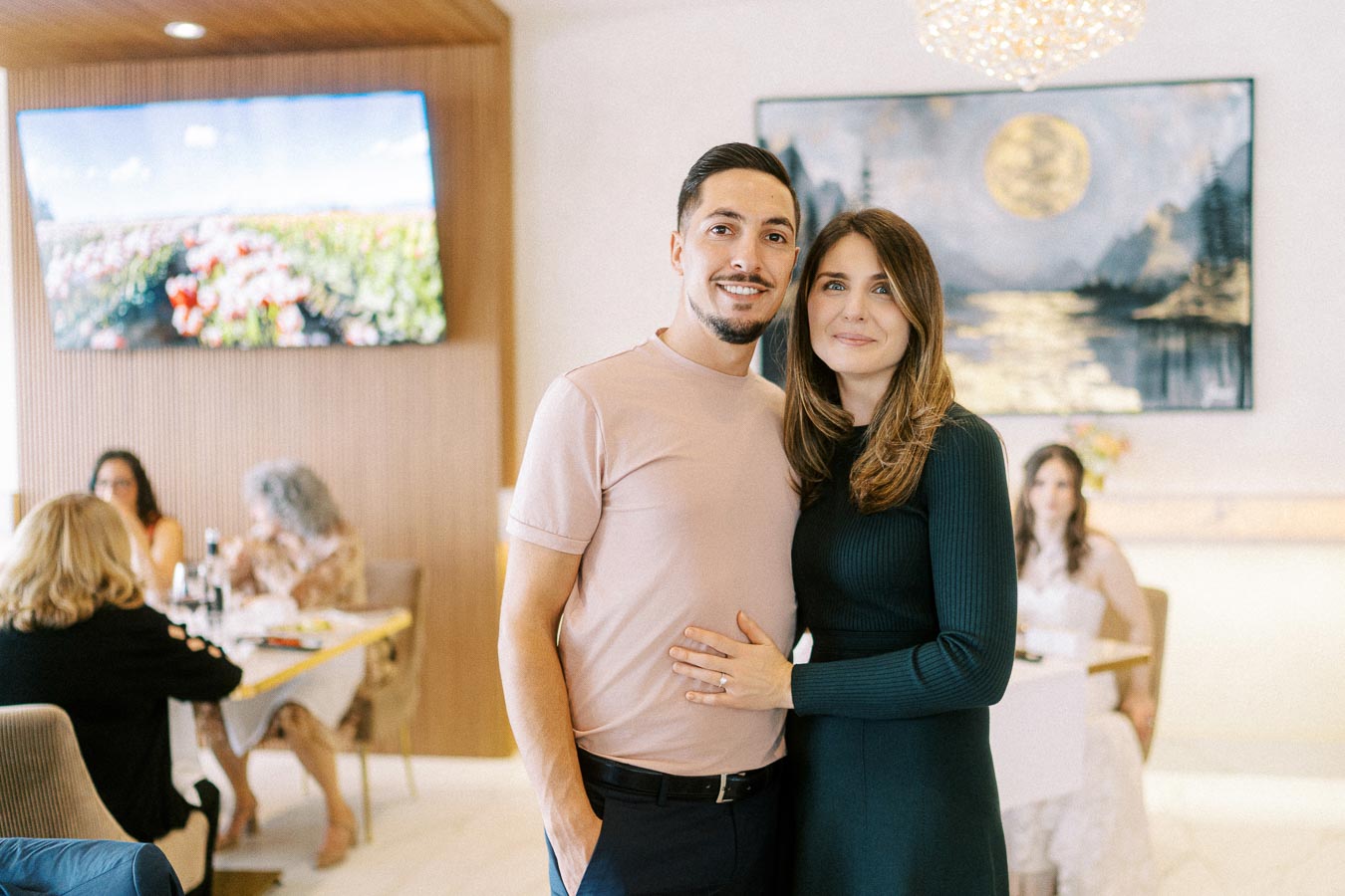 Couple posing together in a restaurant setting, with a scenic painting and television in the background, surrounded by