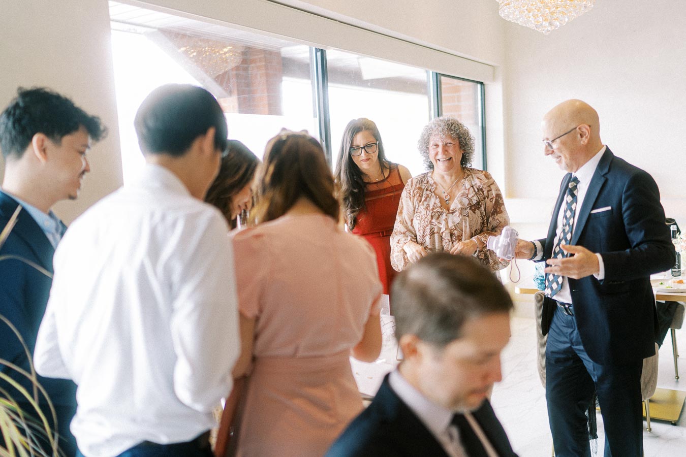 Group of people in formal attire mingling at an indoor event, with a man holding a camera and light streaming through large