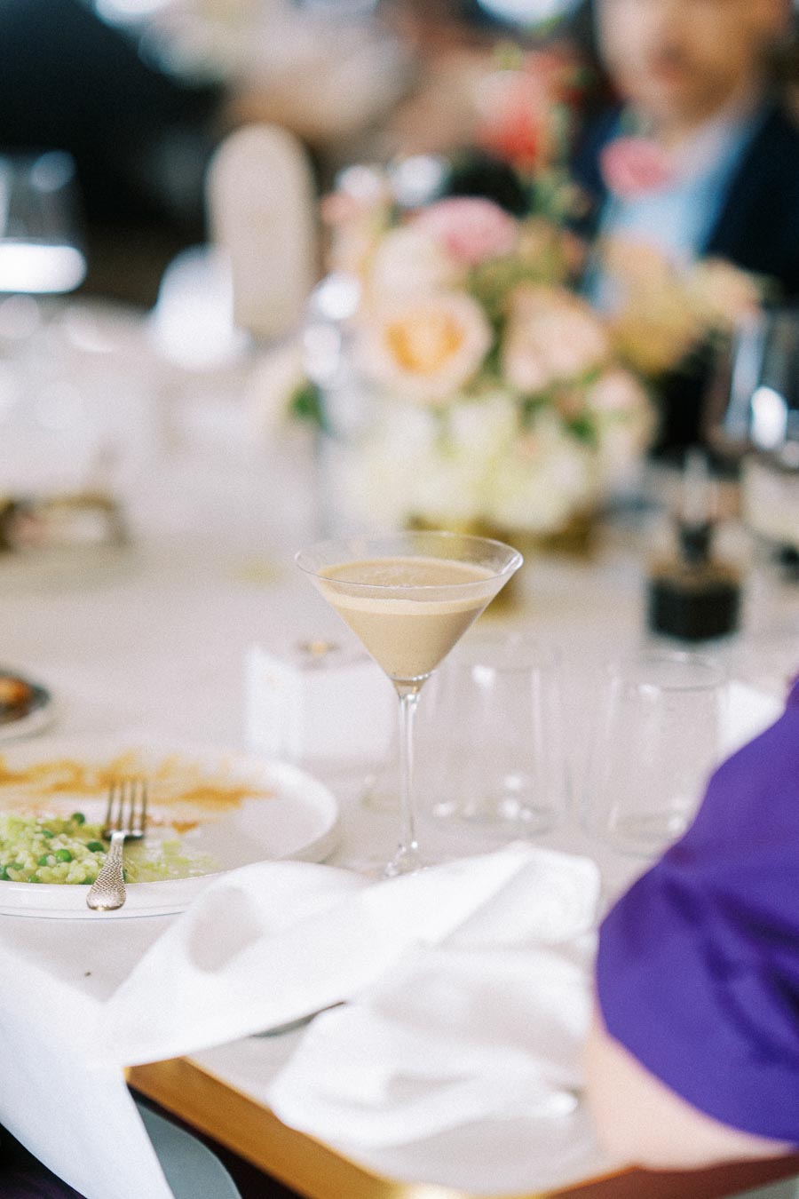 A martini glass with a creamy cocktail on a set table with a plate of food and a floral centerpiece in the background at a