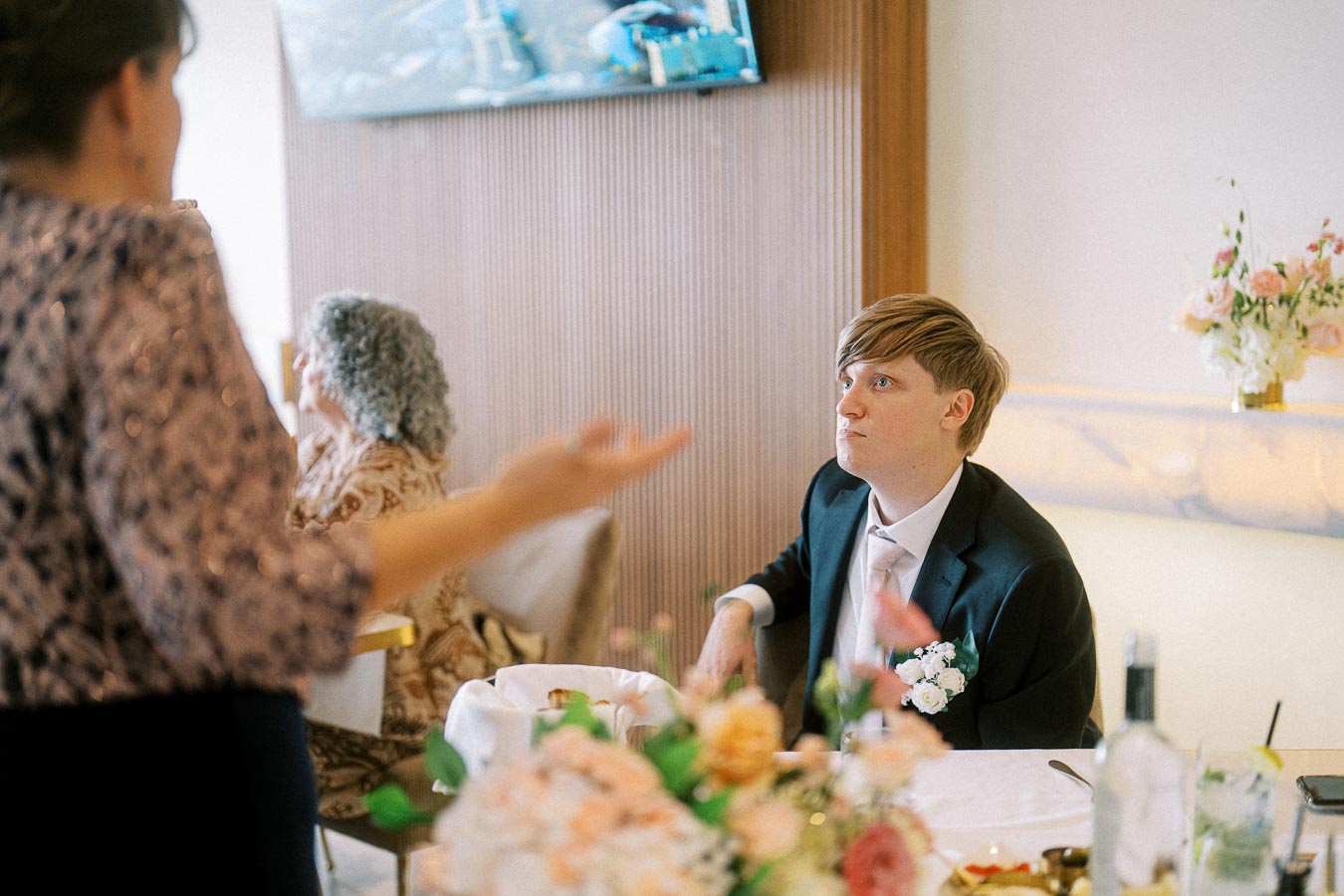 A person in a suit with a floral boutonniere sitting at a dining table, attentively listening during a social gathering,