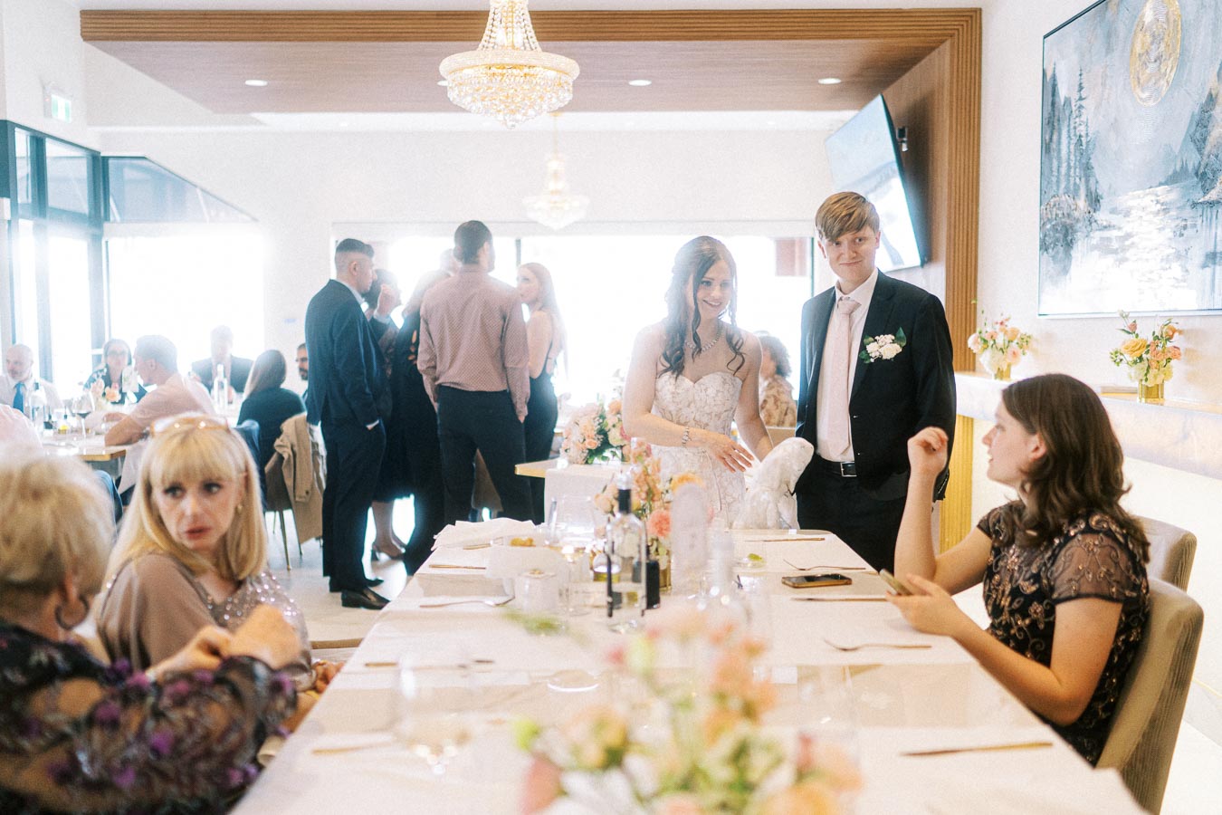 Wedding reception scene in a bright, elegant hall. A bride in a white gown and a groom in a suit stand together near a