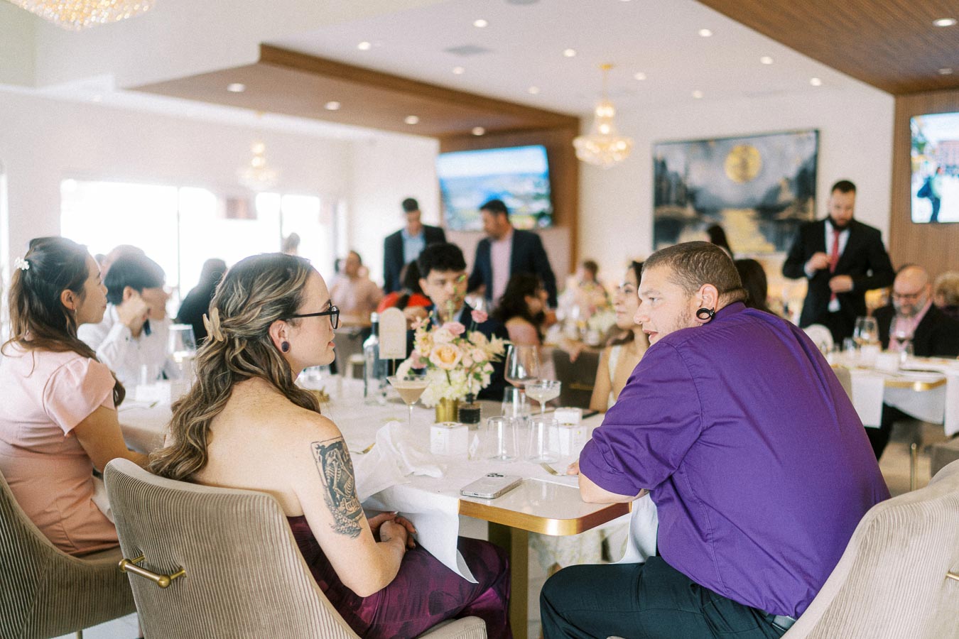 A vibrant gathering at a formal event with elegantly dressed guests seated around a table adorned with floral centerpieces