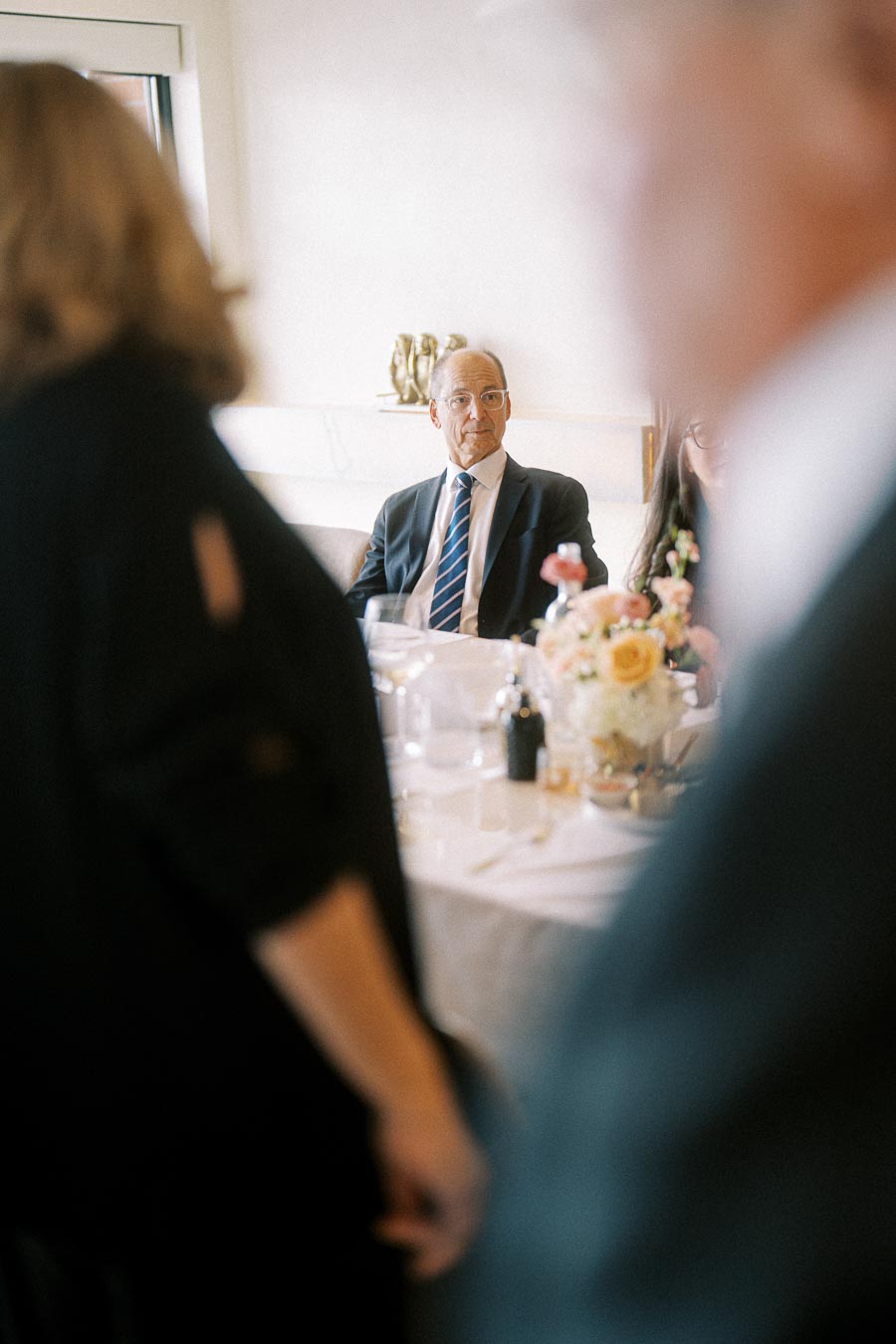 Elegant gathering at a formal event, with a man in a suit sitting at a decorated table, surrounded by people engaged in