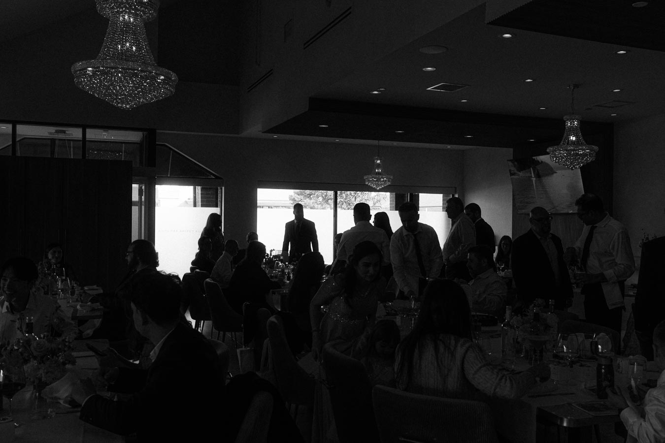 A dimly lit banquet hall filled with people socializing and dining at tables, illuminated by elegant chandeliers.