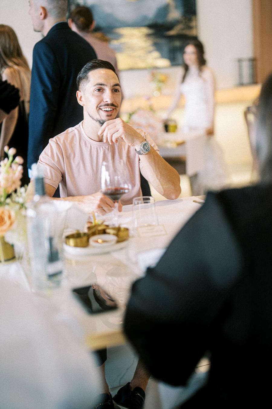 Man in a casual pink shirt smiling and enjoying a glass of red wine at a sophisticated social gathering.