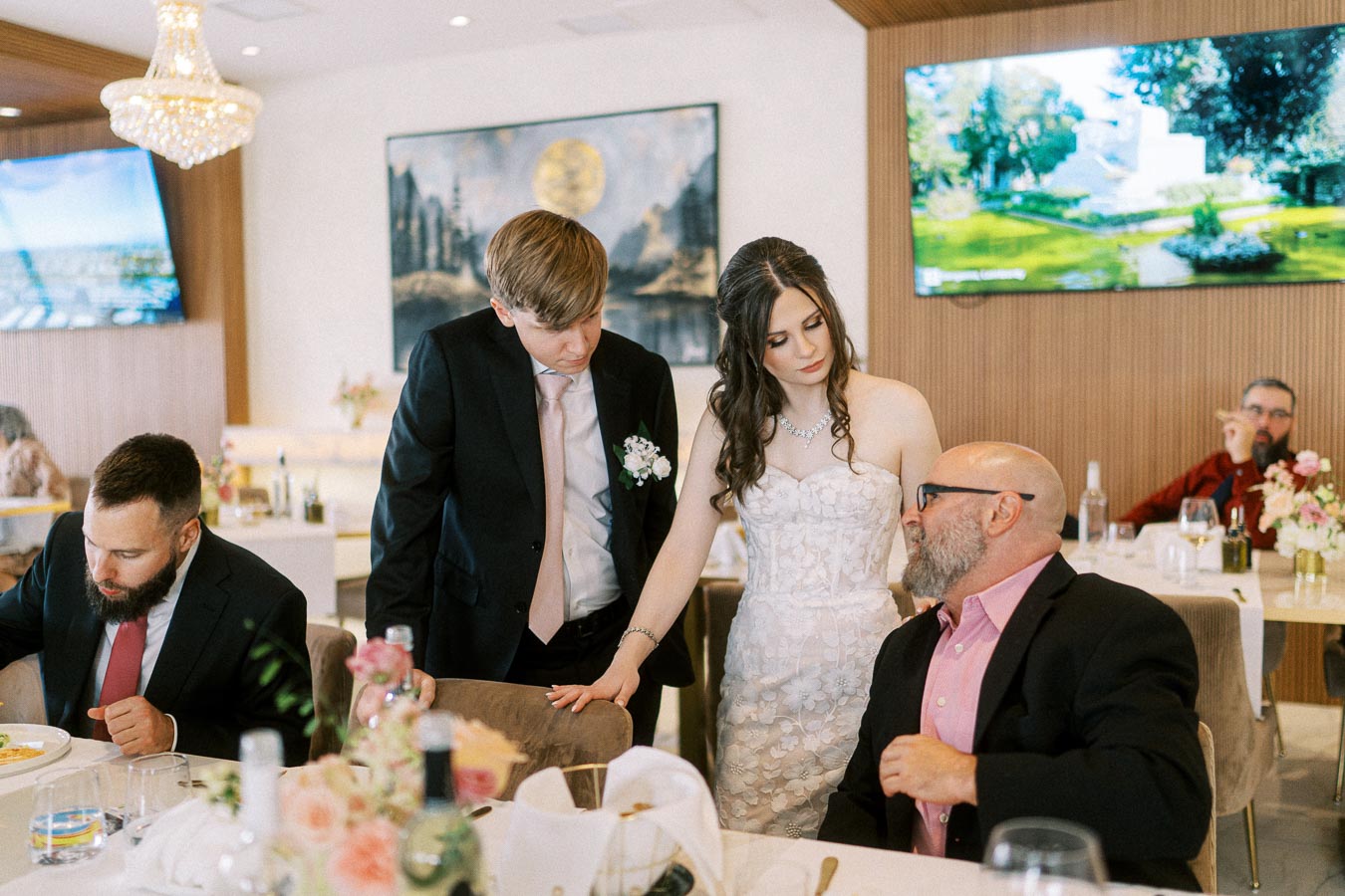A bride in a white wedding dress and a groom in a black suit with a boutonnière interact with seated guests at a reception.