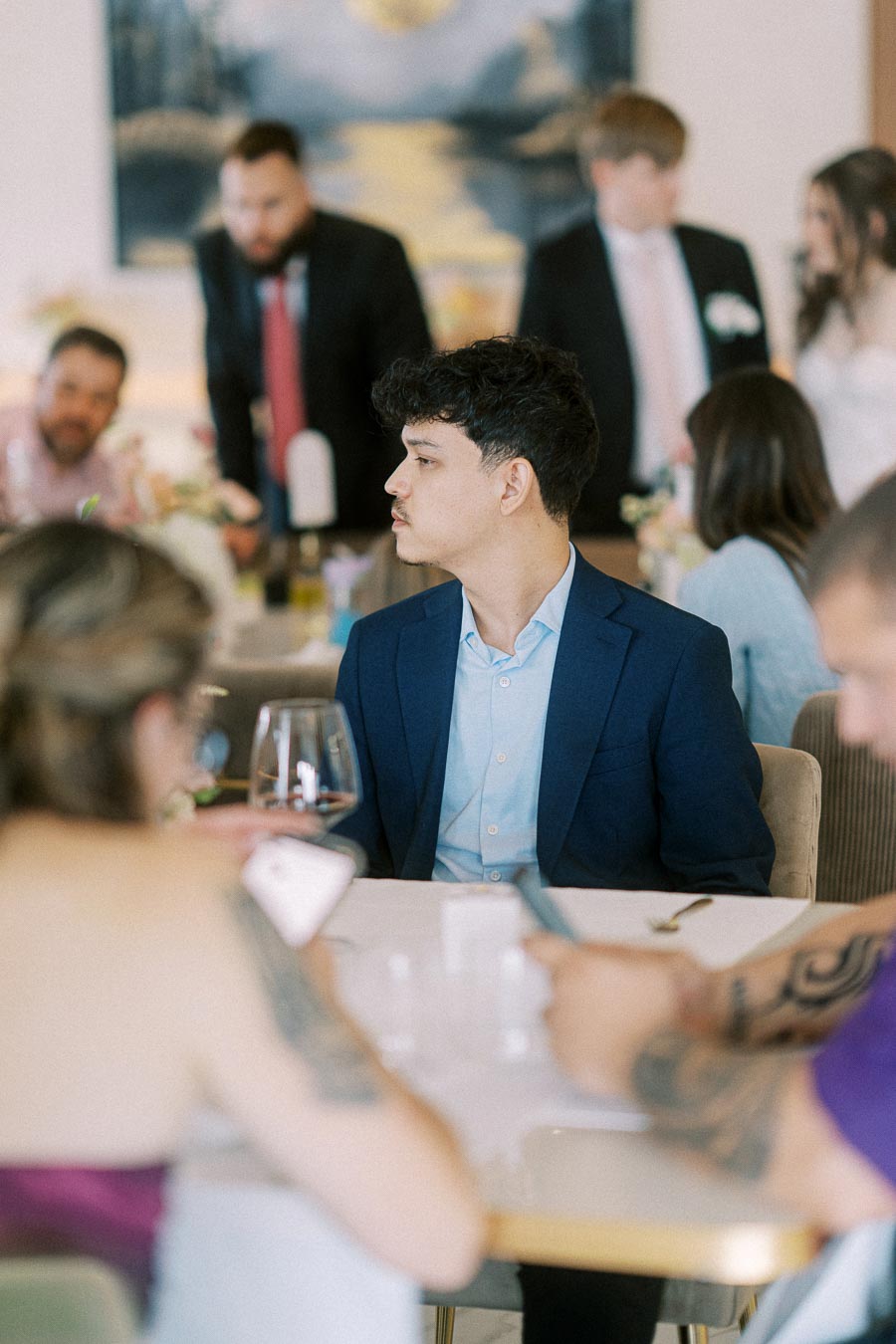 A young man in a blue suit sits at a dining table during a social gathering. People in formal attire engage in conversation