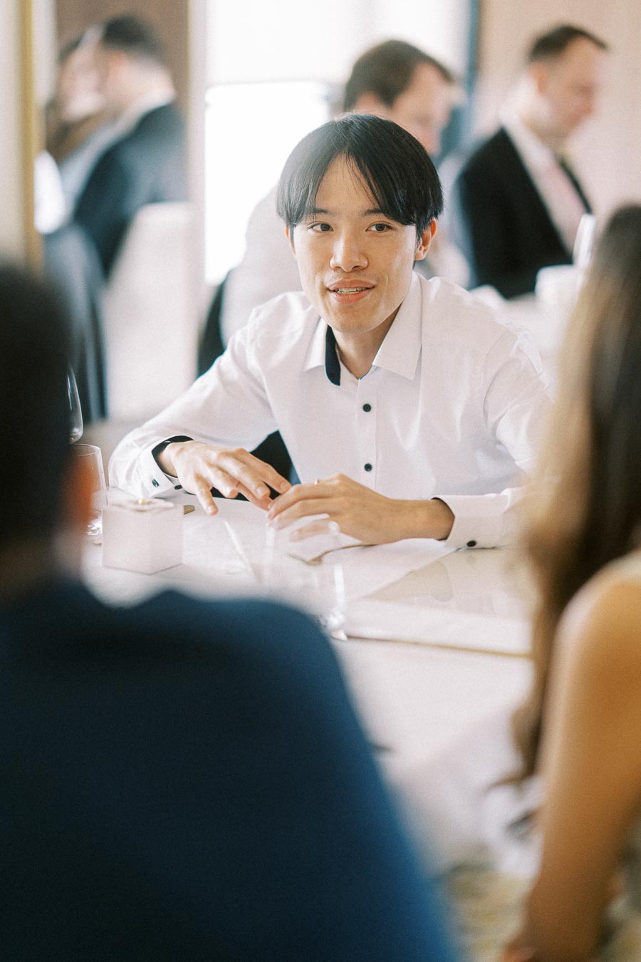 Person in a white shirt engaging in conversation at a formal dinner event, surrounded by elegantly dressed guests.