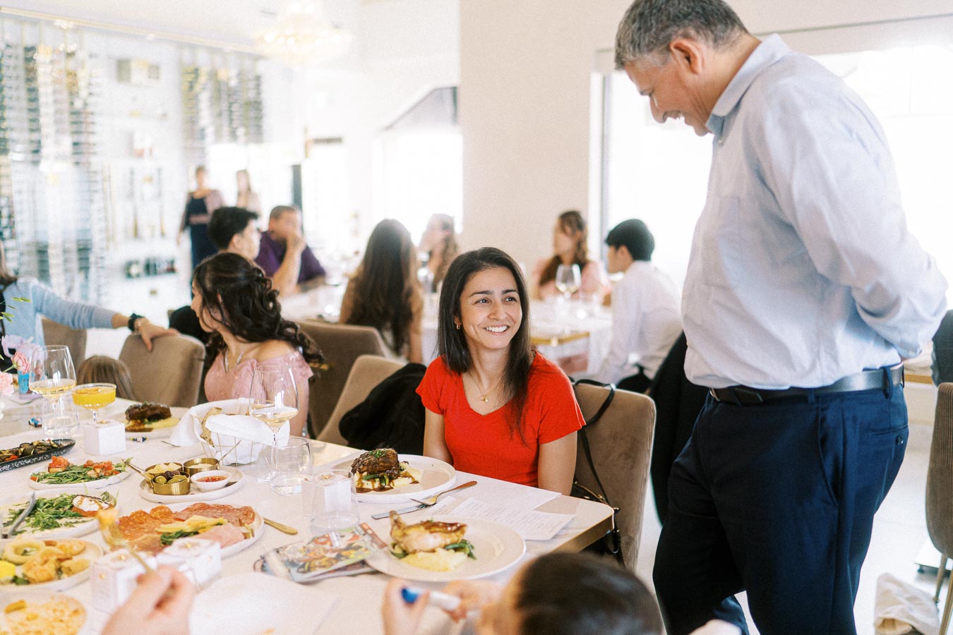 A group of people enjoying a meal at a restaurant, featuring a smiling woman in a red shirt and a man standing beside her.